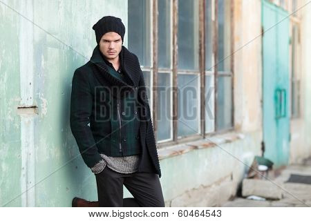 young fashion man posing outdoor, by an old building, with his hands in his pockets and a foot on the wall while looking into the camera