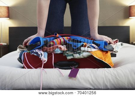 Low section of woman kneeling on overstuffed suitcase in bed