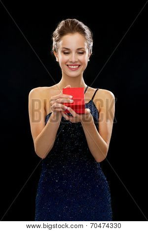 holidays, presents, luxury and happiness concept - smiling woman in dress holding red gift box over black background