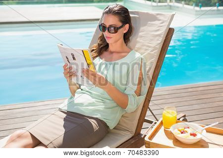 Beautiful young woman reading book by swimming pool with breakfast on table