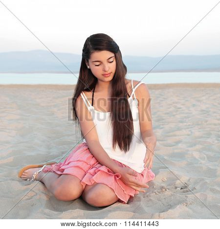 Young causcasian beautiful girl with long dark hair sitting on a beach and playing with sand.