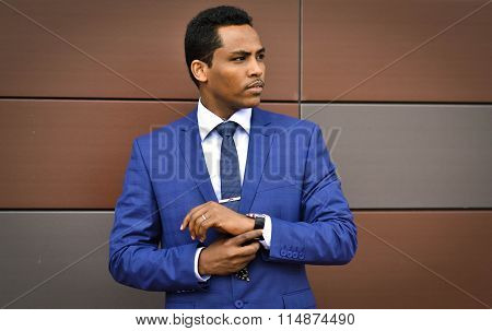 African important,handsome,young person,happy businessman,worker corrects hours on his arm,looks for job or reached career top,stands near the main office.His official style is perfect,face is serious