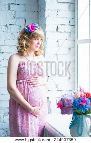 Pregnant woman smiling, looking at belly, holding flowers. Pink dress