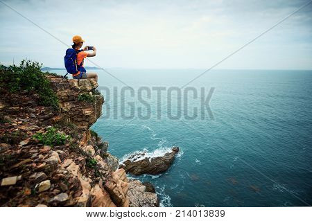 Young woman hiker taking photo with smartphone sit on seaside cliff
