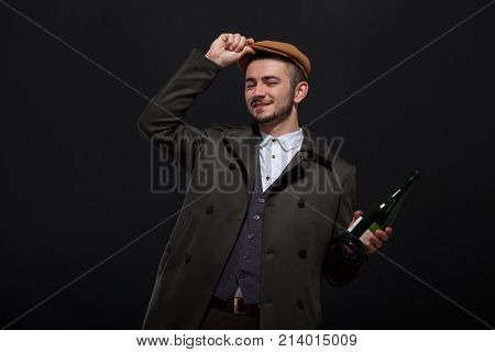 A man on a black background, retro style, in a cap stands with a green bottle of alcohol, hand adjusts the cap