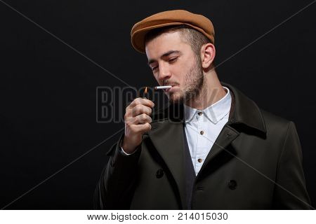 A man on a black background, retro style, in a cap lights a cigarette, screwing up his eyes.