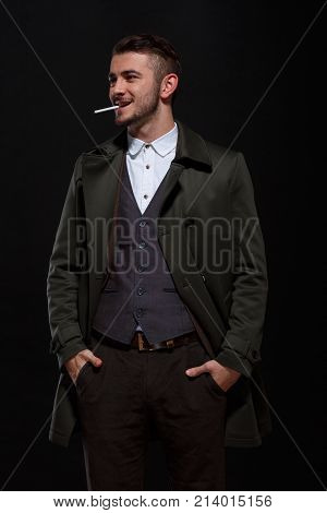 A man on a black background, retro style, stands with a cigarette in his mouth, smiles, putting his hands in his pockets, looking away.
