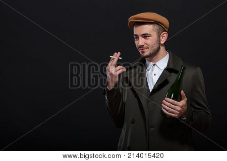 A man on a black background, retro style, holds a bottle of alcohol in his hand, a cigarette sticks out between his fingers, looks into the distance.
