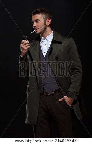 A man on a black background, standing with a cigarette in his mouth, lights up with one hand, a second hand in his pocket