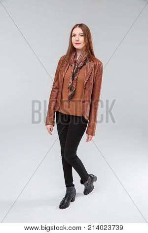 Beautiful brunette with long hair in a brown leather jacket in full growth stands sideways posing in the studio on a gray background.