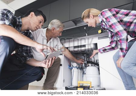 A woman shows plumbers a broken kitchen sink. One of the men inspects the pipes, the second one writes on the tablet. The woman shows the place of breakage.