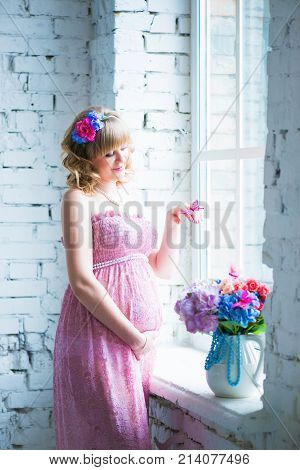 Pregnant woman smiling, looking at belly, holding flowers. Pink dress