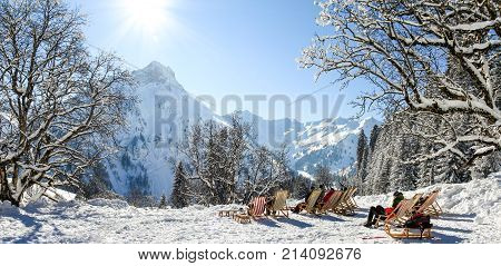 Group of people sitting with deck chairs in sunny winter mountains. Sunbathing in snow. Germany, Bavaria, Allgau, Schwarzenberghuette.