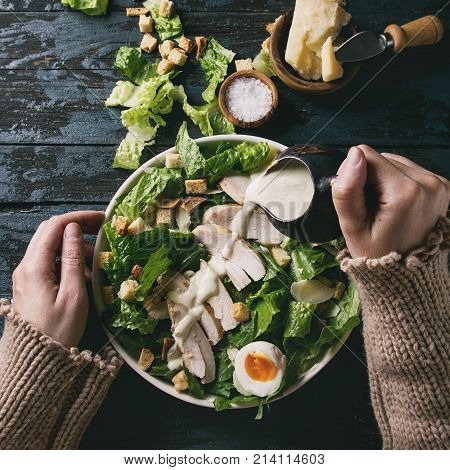 Female hands powring dressing to Classic Caesar salad with chicken breast in white ceramic plate. Served with ingredients above over old dark blue wooden background. Flat lay. Rustic style