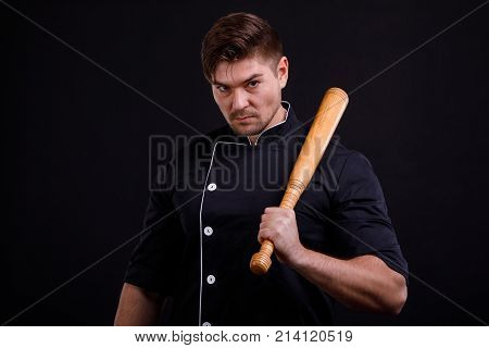 A serious man, pumped into a black tunic, is holding a bat on his shoulder against a black background