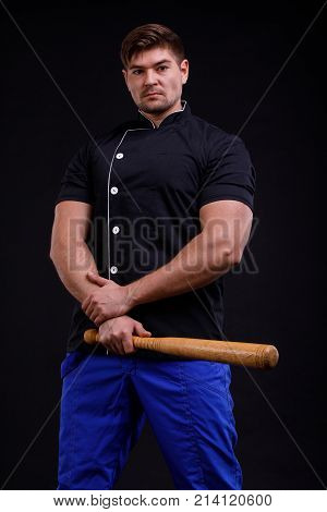 A man with arms crossed holds a bat in a black tunic and blue pants on a black background