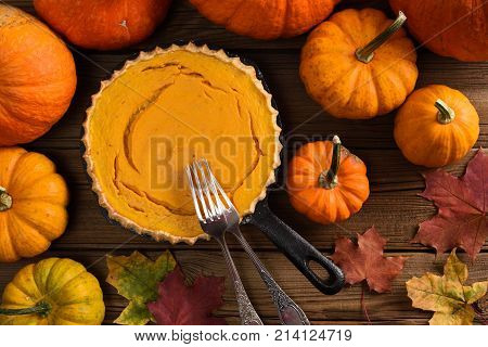 Traditional pumpkin pie flat lay. Open homemade pie baked in cast iron pan surrounded with bright pumpkins and marple leaves on rustic background overhead view