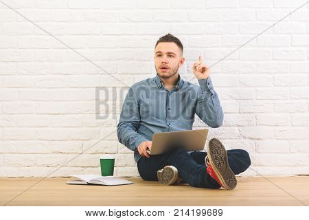 Handsome man sitting in studio with laptop book and coffee cup on wooden floor. White brick wall background. Education idea youth hipster concet