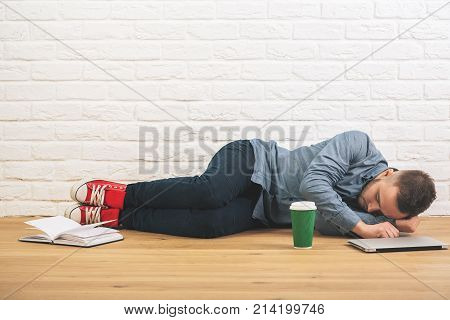 Caucasian guy sleeping in studio with laptop book and coffee cup on wooden floor. White brick wall background. Education tired overworked youth hipster concet