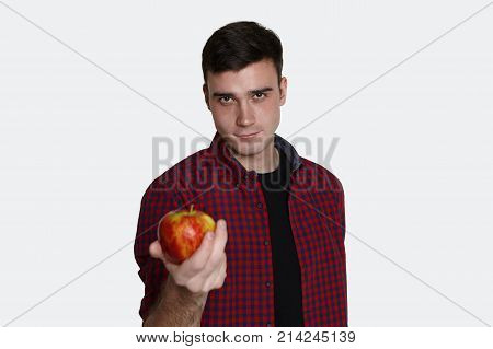 Isolated portrait of confident charming young brunette male dressed casually smiling mysteriously standing in studio with hand reached out at camera offering you big mellow apple. Selective focus