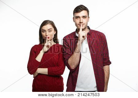 Attractive young Caucasian couple pretty woman and handsome man asking for silence keeping index fingers on their lips saying Shh. People relationships secrecy and confidential information