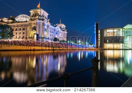 BERLIN GERMANY - AUGUST 26 2017; Crowd seated along embankment in front Riechstag building for night light and picture show across River Spree illuminated buildings and reflected colors on calm water under dark sky.