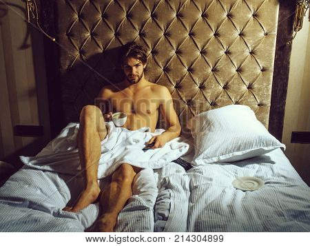 young handsome bearded man with bare muscular chest on sexy body in white bedsheet laying on bed in hotel bedroom with remote control and coffee cup