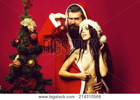 bearded santa claus man with long beard in new year coat with pretty sexy naked girl or woman in hat near decorated Christmas tree on pink background