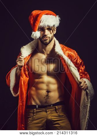 Handsome santa claus guy sexy young bearded macho christmas man model in red xmas or new year coat and hat has bare muscular torso and chest on black background