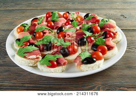 Horizontal shot of a plate with canapes with ham cherry tomatoes and black olives decorated with rucola rucoli plant edible vegetables bacon jamon appetizers menu restaurant.