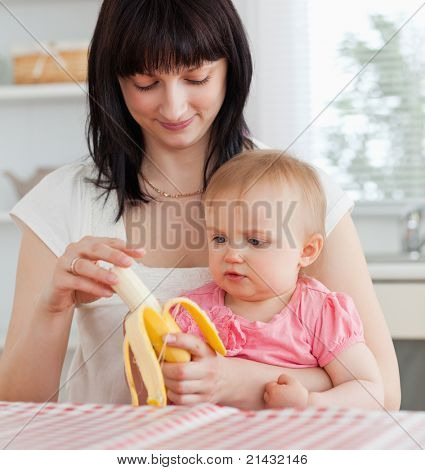 Okouzlující bruneta žena žáčkům banán současně držet své dítě na kolenou v kuchyni