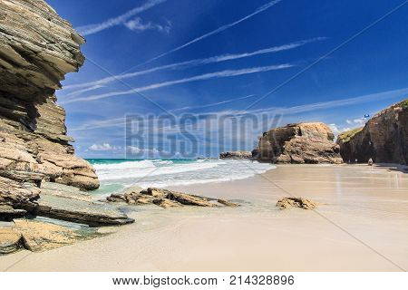 View of the ocean between rocks of Praia de Augas Santas. A fragment of the beach of cathedrals in summer day in good weather. The blue sky with numerous traces of planes