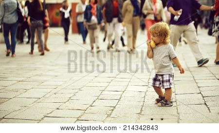 Photo closeup of cute fair-haired blond kid tiny little child baby boy eating bun standing on flag-stone pavement in crowd cityscape on blurred grey background horizontal picture