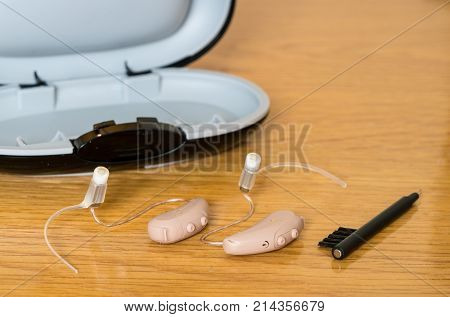 Macro close up of a matched pair of tiny modern hearing aids with case and brush on wooden bedside dressing table