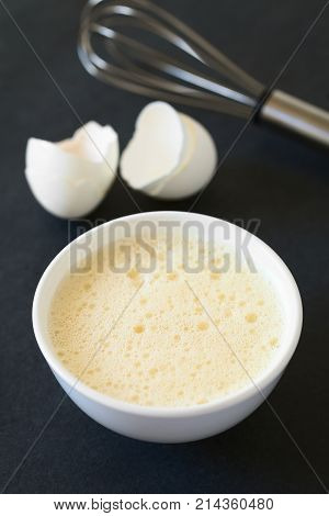 Whisked whole eggs (whites and yolks together) in bowl with whisk and egg shells in the back photographed on slate with natural light (Selective Focus Focus in the middle of the whisked egg)