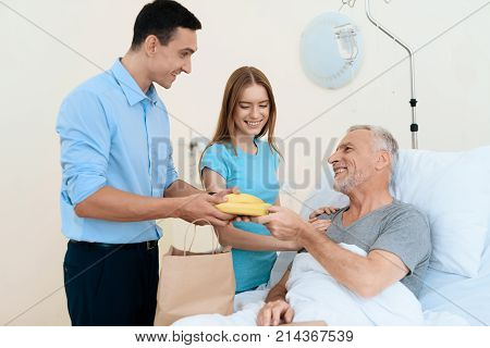 An elderly man lies in a hospital room on a bed. He is seen by a man with a woman. A woman is standing next to him. A man gives an old man a banana and that one is struck.