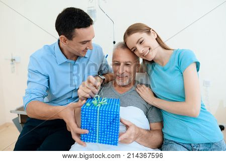 An elderly man lies in a hospital room on a bed. He is seen by a man with a woman. They brought the old man a gift in a large box and hug him. The old man smiles.