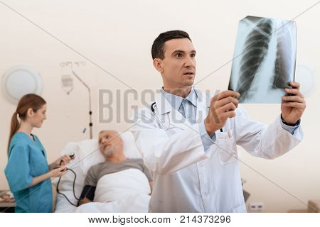 The old man lies on a cot in the medical ward, and next to him there is a doctor and a nurse. The doctor is holding an X-ray in his hands, the nurse is standing next to him. They are in modern clinic.