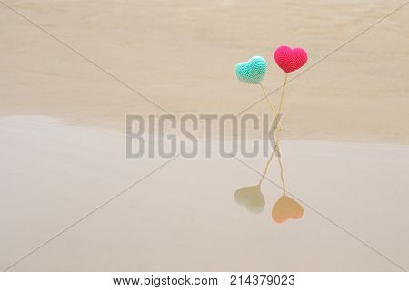 Two crocheted bright and colorful hearts on wooden sticks on beach reflected in water. Pink red and blue turquoise hearts. Texture of sand. St. Valentine's Day theme. Love and wedding theme