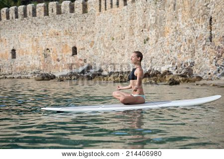 Fit woman in swimwear doing meditating on stand up paddle with medieval castle wall behind her