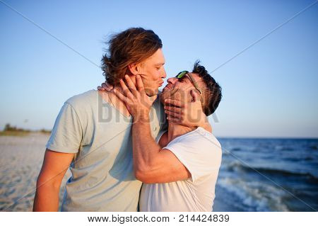 Two men embrace against the background of the sea. They represent a playful kiss.