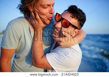 Two men embrace against the background of the sea. They represent a playful kiss.
