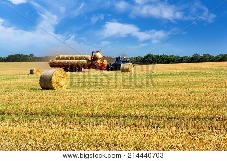Tractor carrying hay at field in summer day. Landscape with a field of straw rolls sky and clouds.