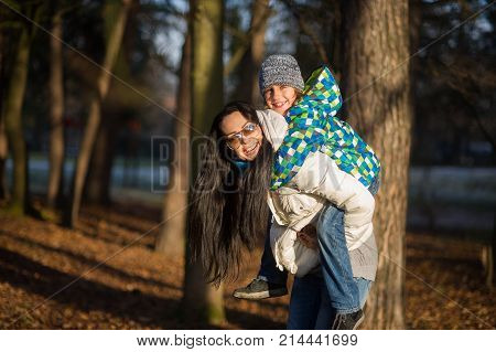 Young woman bears on a back of the boy of 9-10 years. Mum and the son on walk in autumn park. Serene autumn day good mood.