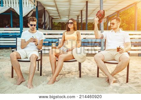 Lifestyle Young people enjoying summer vacation sunbathing drinking at beach bar. Two guys and a young girl sit on the terrace of the summer bar and enjoy the conversation Sun flare
