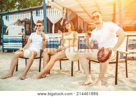 Lifestyle Young people enjoying summer vacation sunbathing drinking at beach bar. Two guys and a young girl sit on the terrace of the summer bar and enjoy the conversation Sun flare