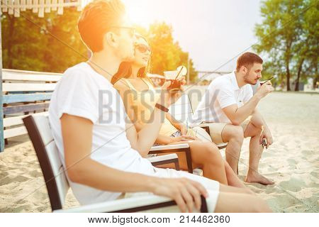 Lifestyle Young people enjoying summer vacation sunbathing drinking at beach bar. Two guys and a young girl sit on the terrace of the summer bar and enjoy the conversation Sun flare