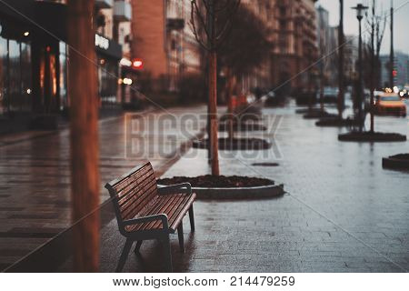 Empty wet lonely bench standing on pavement boulevard in rainy autumn day with raws of naked trees around cars houses and illuminated signboards in defocused background with strong bokeh evening