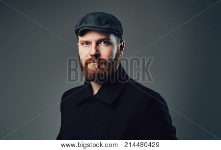 Portrait of Redbeard elegant male dressed in a tweed flat cap and a black jacket over grey background.