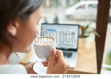 Rearview close up shot of a woman halding a cup of fresh coffee sitting at the cafe with her laptop in front of the window looking out at the street urban lifestyle relaxation businesswoman copyspace.